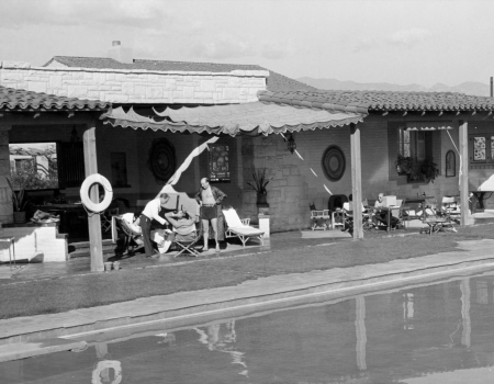 In a black-and-white courtyard, people lounge by a pool in front of a stone house with awnings and garden chairs.