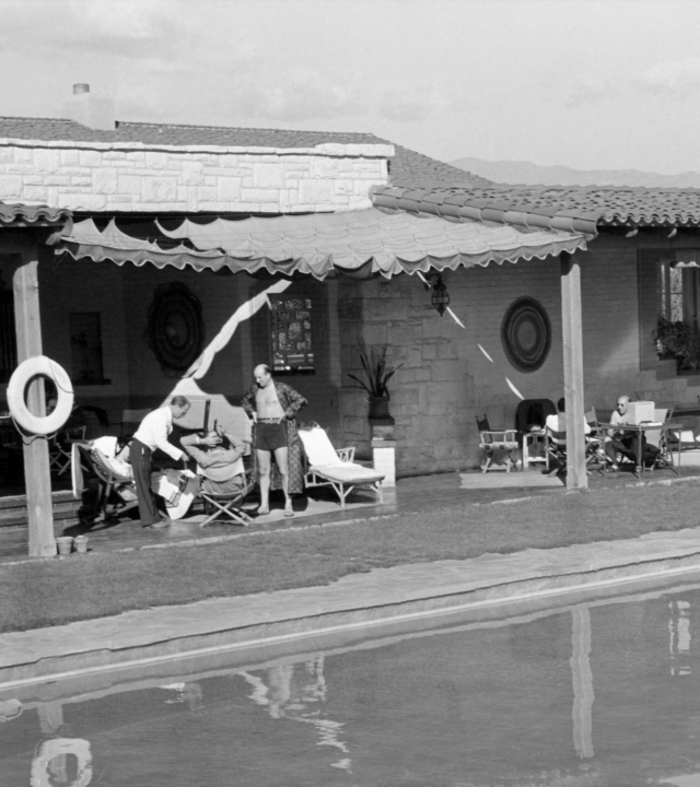 In a black-and-white courtyard, people lounge by a pool in front of a stone house with awnings and garden chairs.