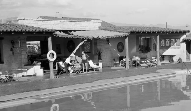 In a black-and-white courtyard, people lounge by a pool in front of a stone house with awnings and garden chairs.