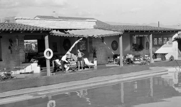 In a black-and-white courtyard, people lounge by a pool in front of a stone house with awnings and garden chairs.