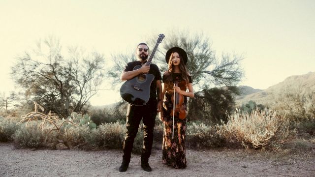 A man with a guitar and a woman with a violin stand in a desert landscape, dressed in casual, bohemian outfits, overcast sky.
