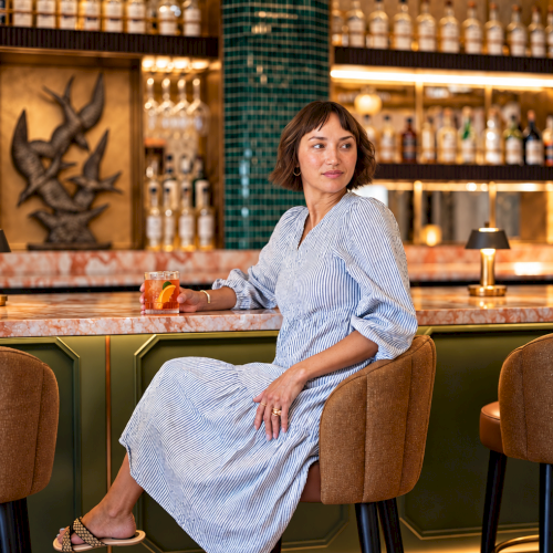 A woman in a striped dress sits at a bar, holding a cocktail with orange and ice, surrounded by bottles and warm lights.