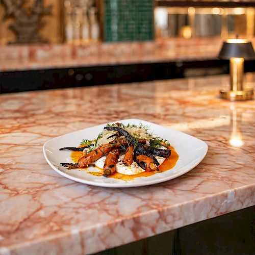 A plated gourmet meal of shrimp and vegetables with herbs sits on a marble table in a warmly lit restaurant.