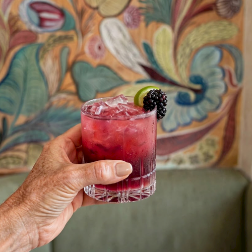 A hand holds a chilled pink cocktail in a glass with ice, topped with a lime slice and blackberries against graffiti.