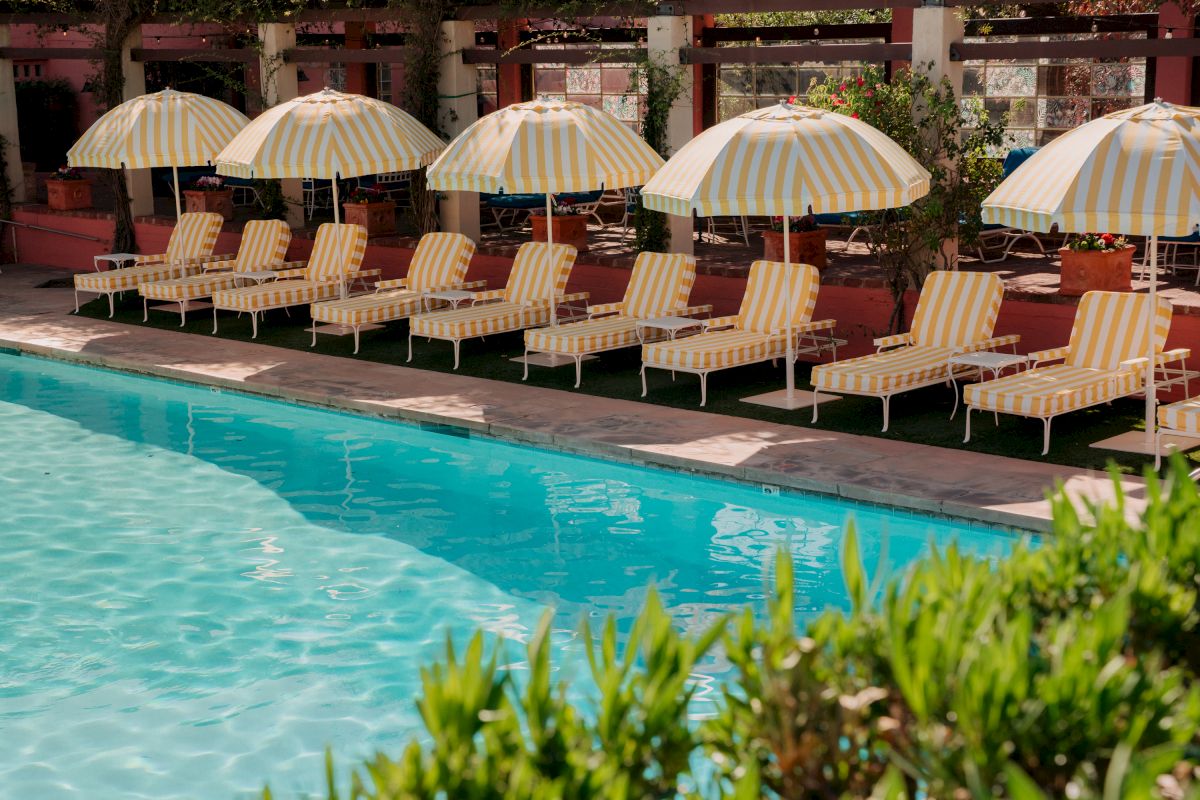 An outdoor poolside lounge with yellow-and-white striped umbrellas and striped chairs, surrounded by plants and warm sunlight, and a swimming pool in front.