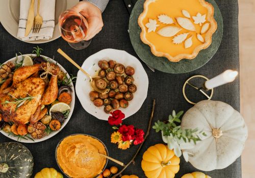 A festive spread of plated food: roasted chicken, mushrooms, olives, a veggie tart, wine, bread, pumpkin and flowers on a table.