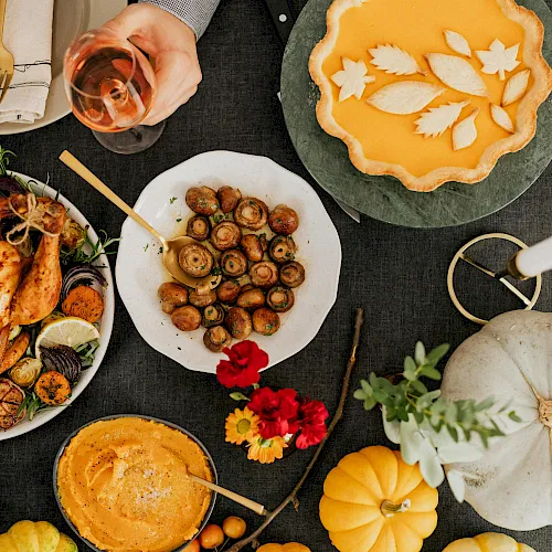 A festive spread of plated food: roasted chicken, mushrooms, olives, a veggie tart, wine, bread, pumpkin and flowers on a table.