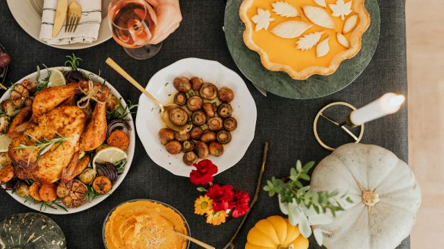 A festive spread of plated food: roasted chicken, mushrooms, olives, a veggie tart, wine, bread, pumpkin and flowers on a table.
