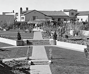 A black-and-white courtyard garden with a tiled walkway, lush plants and cacti, and stucco buildings with terracotta-tile roofs.