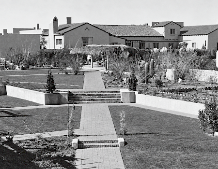 A black-and-white courtyard garden with a tiled walkway, lush plants and cacti, and stucco buildings with terracotta-tile roofs.