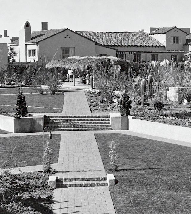 A black-and-white courtyard garden with a tiled walkway, lush plants and cacti, and stucco buildings with terracotta-tile roofs.