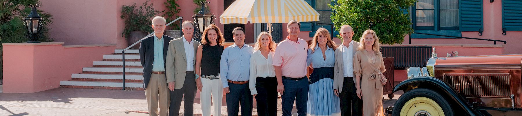 A group poses for a photo outside the Arizona Inn, in front of a pink building, with an umbrella and vintage car nearby.