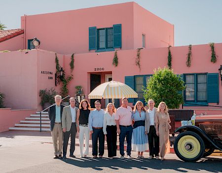 A group poses for a photo outside the Arizona Inn, in front of a pink building, with an umbrella and vintage car nearby.