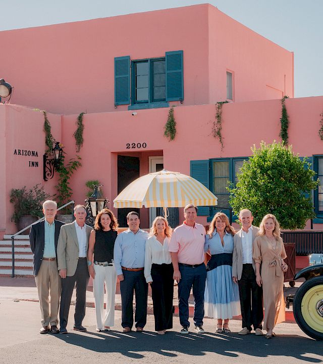 A group poses for a photo outside the Arizona Inn, in front of a pink building, with an umbrella and vintage car nearby.