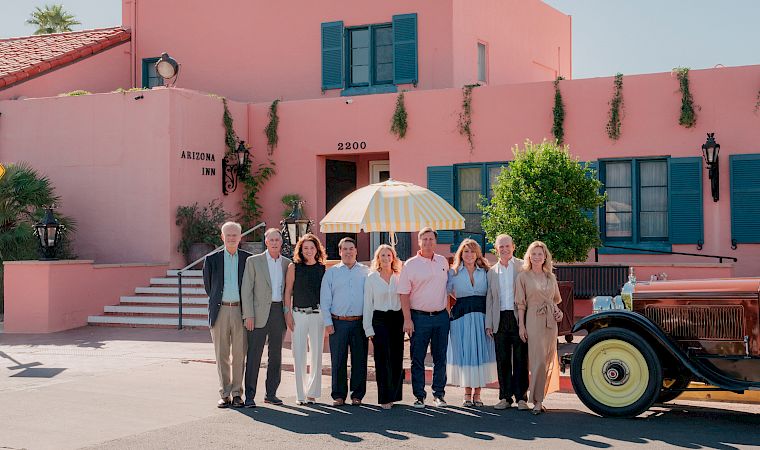 A group poses for a photo outside the Arizona Inn, in front of a pink building, with an umbrella and vintage car nearby.
