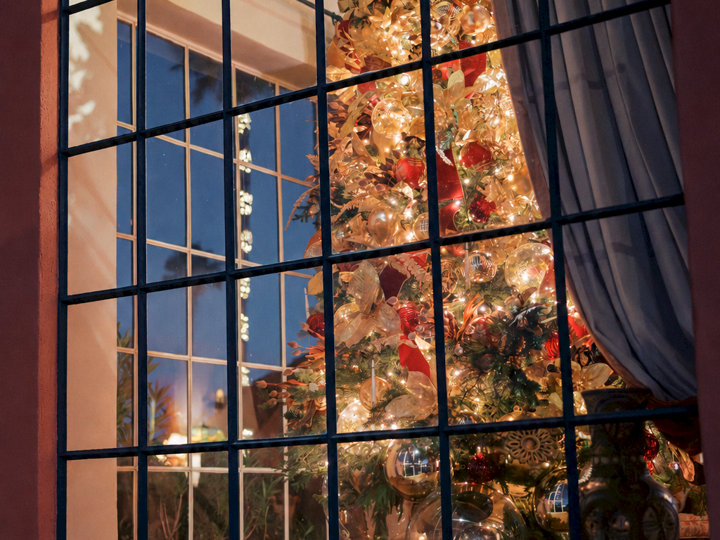 View through a window grid shows a lit Christmas tree wrapped in shiny baubles and lights, with curtains drawn.