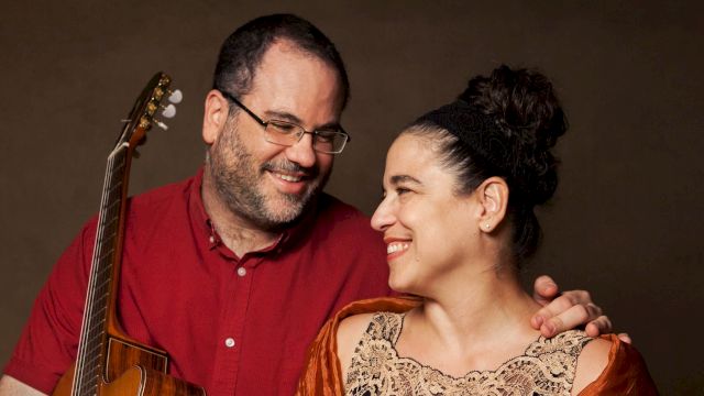 A smiling couple is posing together; the man holds an acoustic guitar while the woman looks lovingly at him.