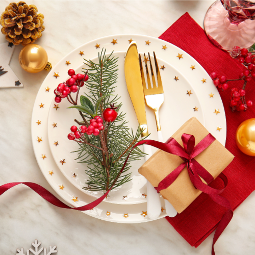 A festive Christmas place setting with a decorative plate, fork and knife, pine sprig and red berries, plus ornaments and gifts laid out.