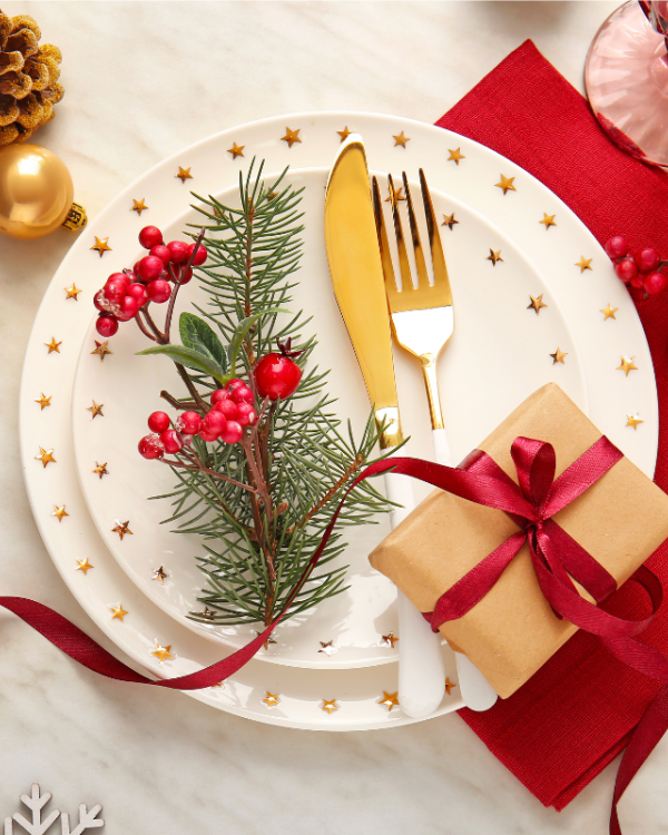 A festive Christmas place setting with a decorative plate, fork and knife, pine sprig and red berries, plus ornaments and gifts laid out.