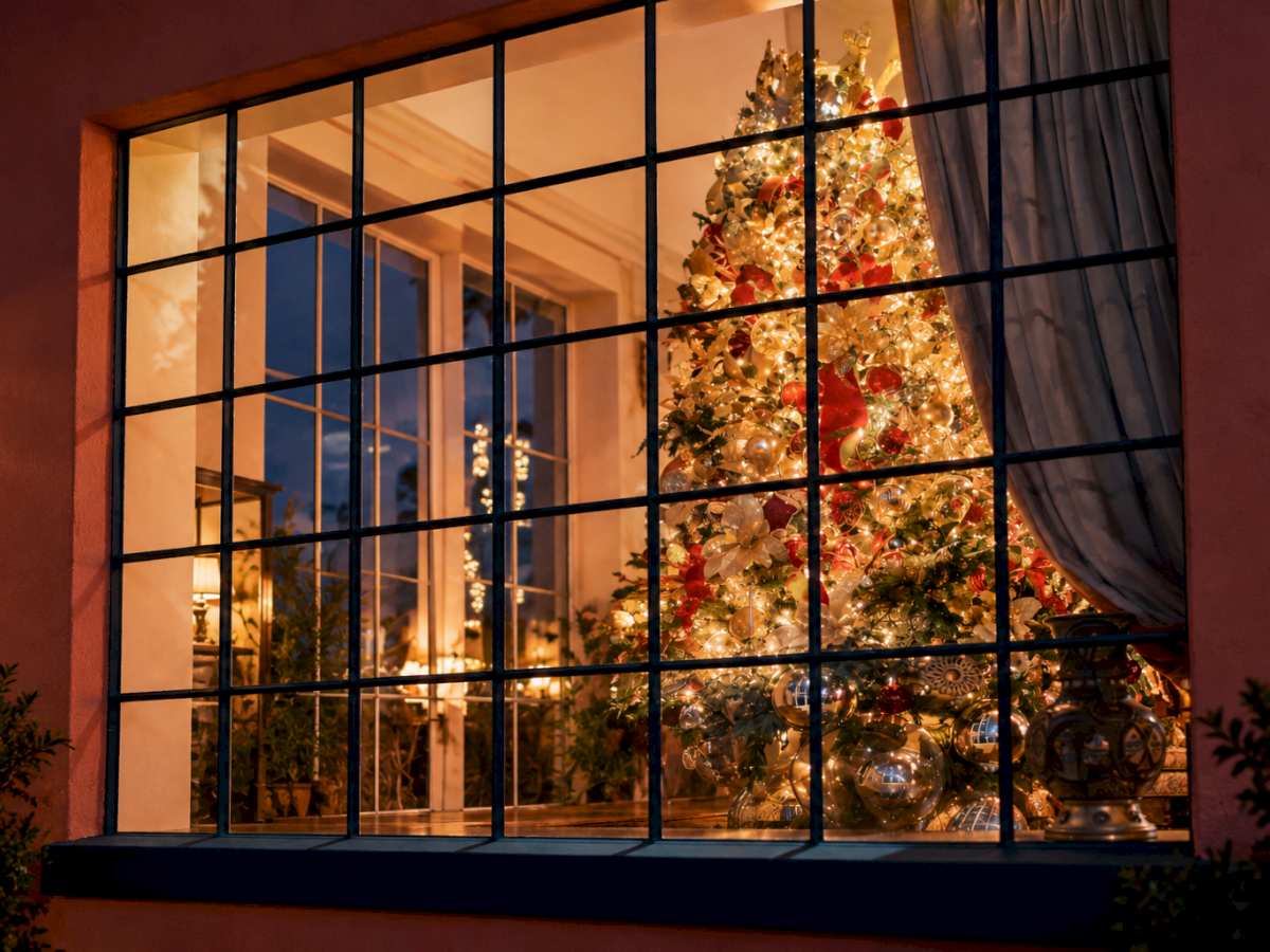A warmly lit room through a window, with a decorated Christmas tree and glowing ornaments inside.