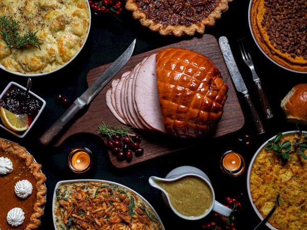 A festive dinner spread with glazed ham, sliced pork, roast turkey, pies, casseroles, and gravy on a dark table.
