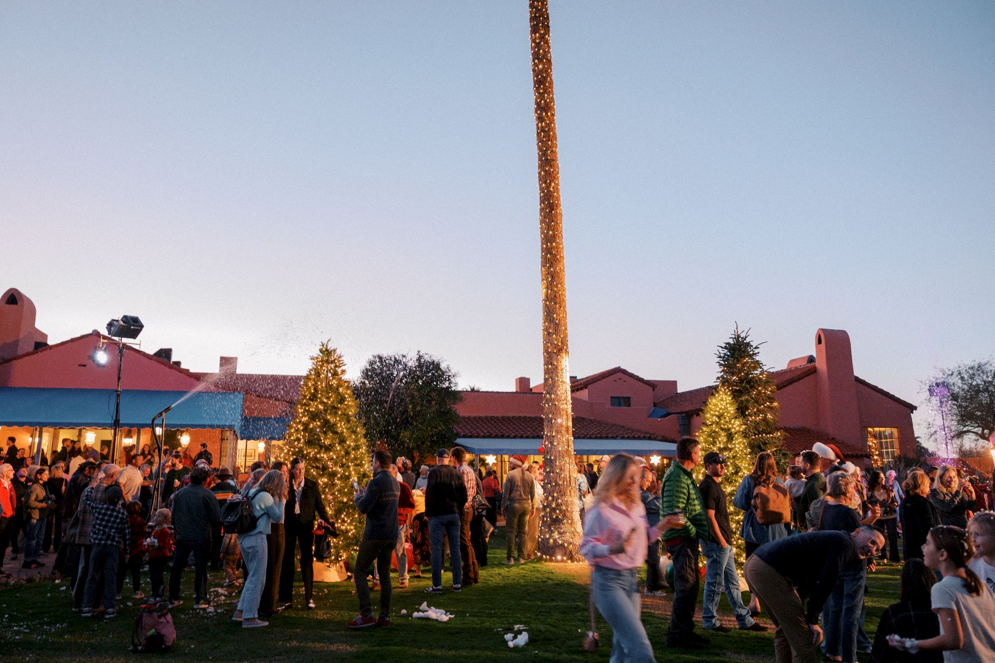 Crowd at a festive outdoor holiday market, with sparkling Christmas trees and a tall lighted pole glowing in the evening.