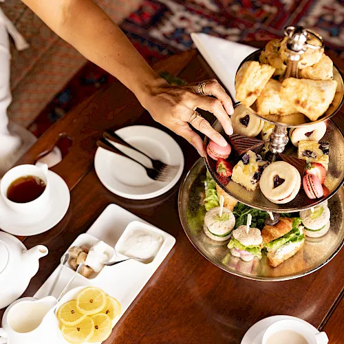 A woman serves afternoon tea on a tiered tray&mdash;mini sandwiches, pastries, and fruit&mdash;along with teacups and teapot on a table.