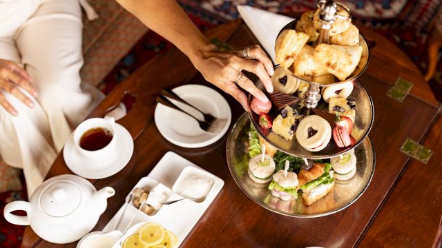 A woman serves afternoon tea on a tiered tray&mdash;mini sandwiches, pastries, and fruit&mdash;along with teacups and teapot on a table.