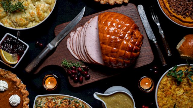 A festive meal spread with glazed ham slices, roast meat, and savory pies with bowls of sides, herbs, and cranberry garnishes.