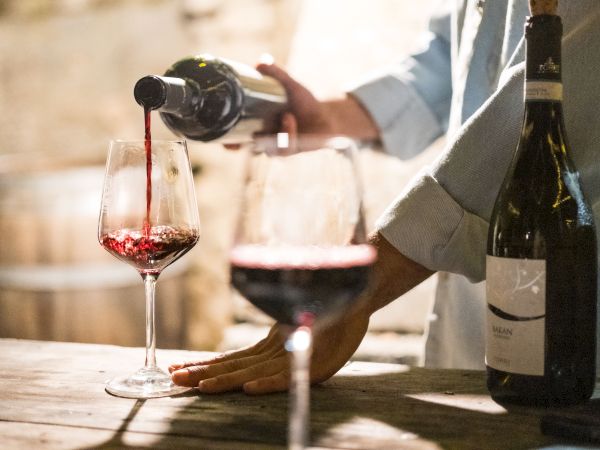 A wine bottle pours red wine into a glass while people dine in a warm, softly lit setting.