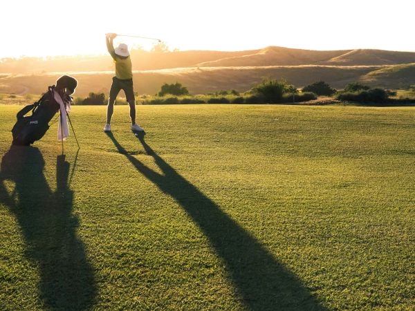 A golfer swings at sunrise on a grassy course as a photographer captures the moment in open hills.