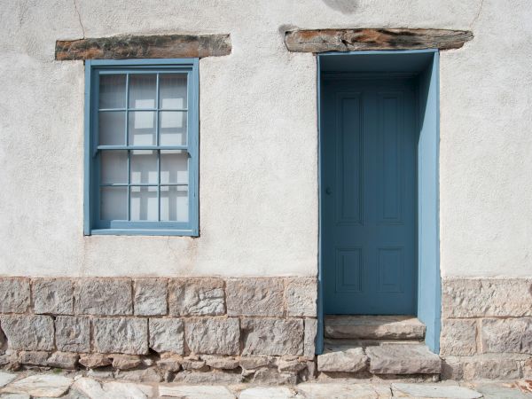 A blue double-door and window sit in a white stucco wall above a stone foundation, with weathered wooden trim overhead.