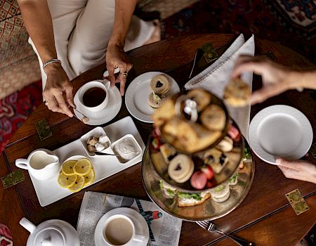 A group breakfast at a wooden table with teacups, pastries/danishes, and people serving coffee and sweets.