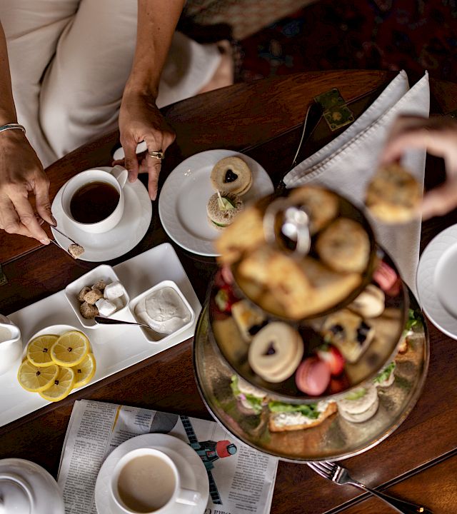 A group breakfast at a wooden table with teacups, pastries/danishes, and people serving coffee and sweets.
