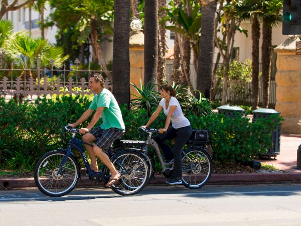 A man and woman ride electric bikes along a sunny garden path lined with palms and greenery.