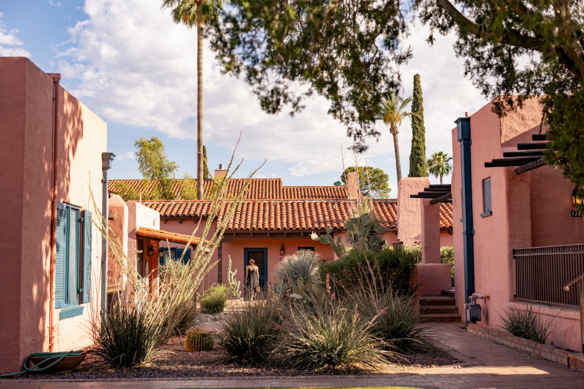 A sunny courtyard with pink stucco buildings, terracotta tile rooftops, tall palms, desert plants, and a clear blue sky.