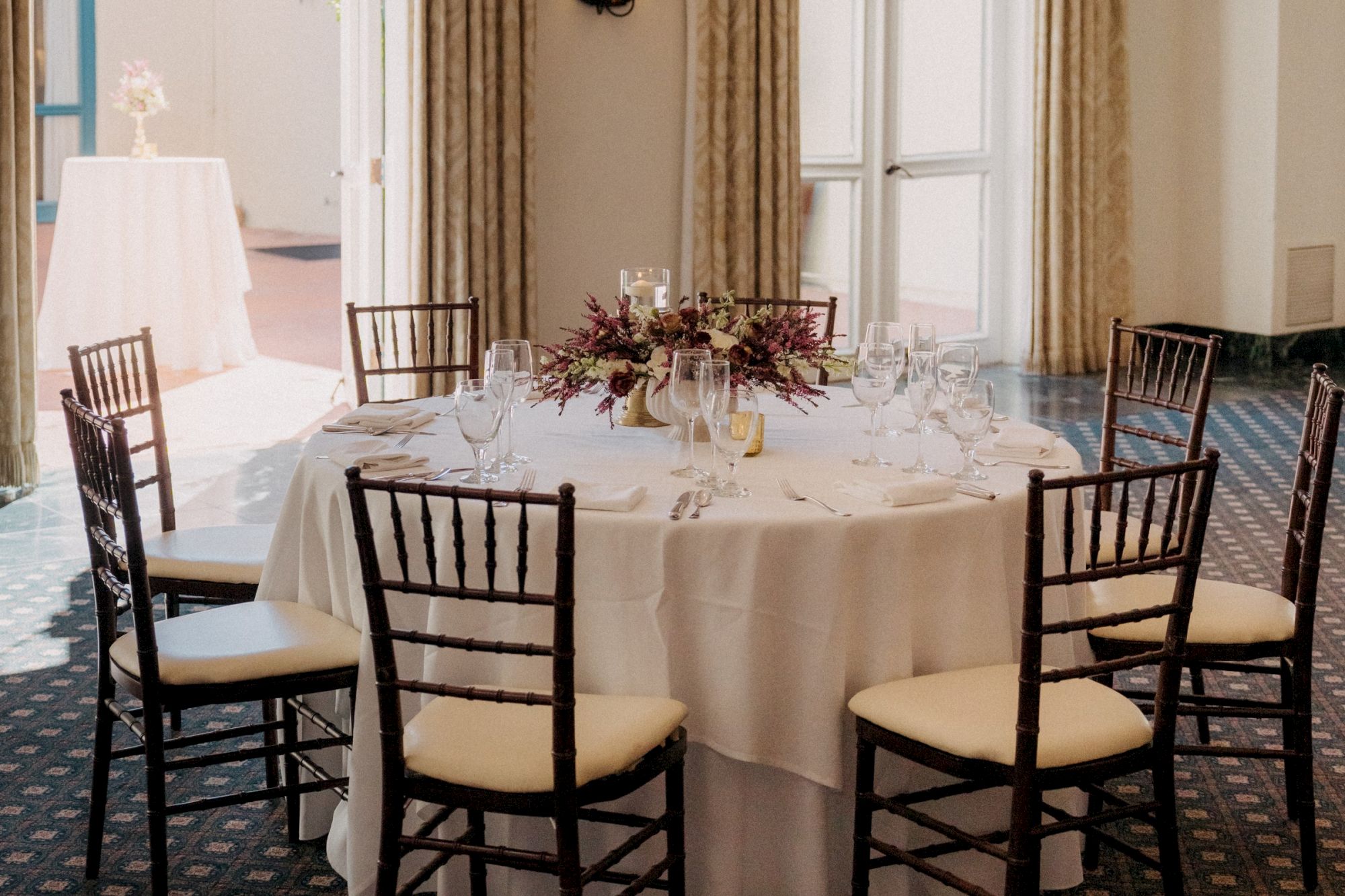 A round banquet table set with white tablecloth, glassware, candles, and dark wood chairs in a formal dining room, ready for a meal.