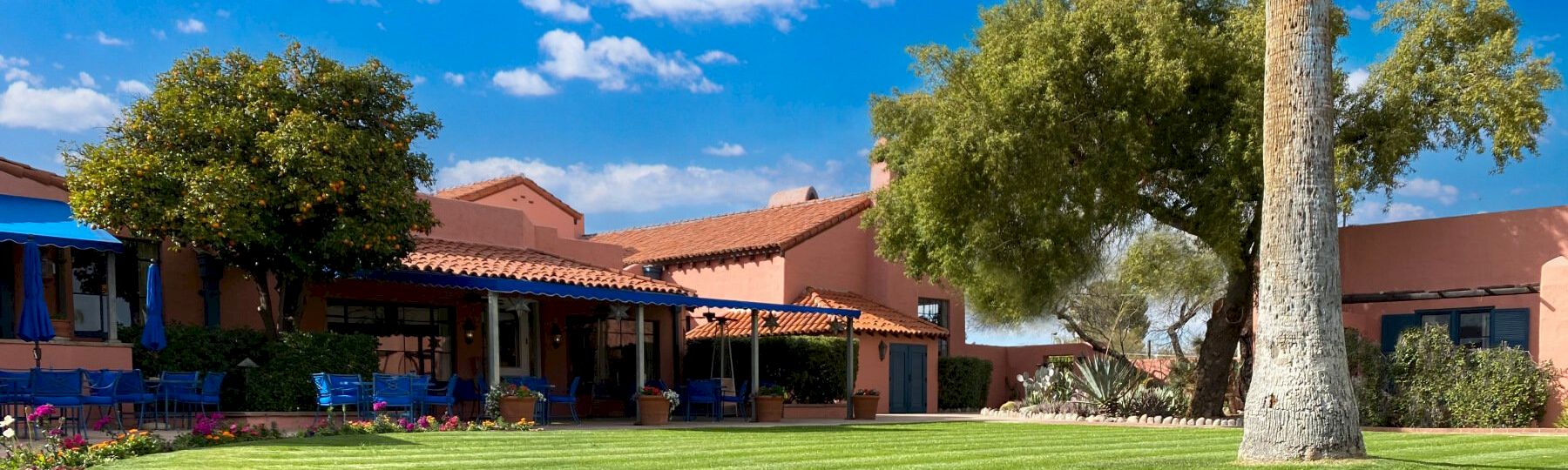 A sunny courtyard with orange-tiled buildings, blue awnings, green lawn, palm trees, and outdoor seating under umbrellas.