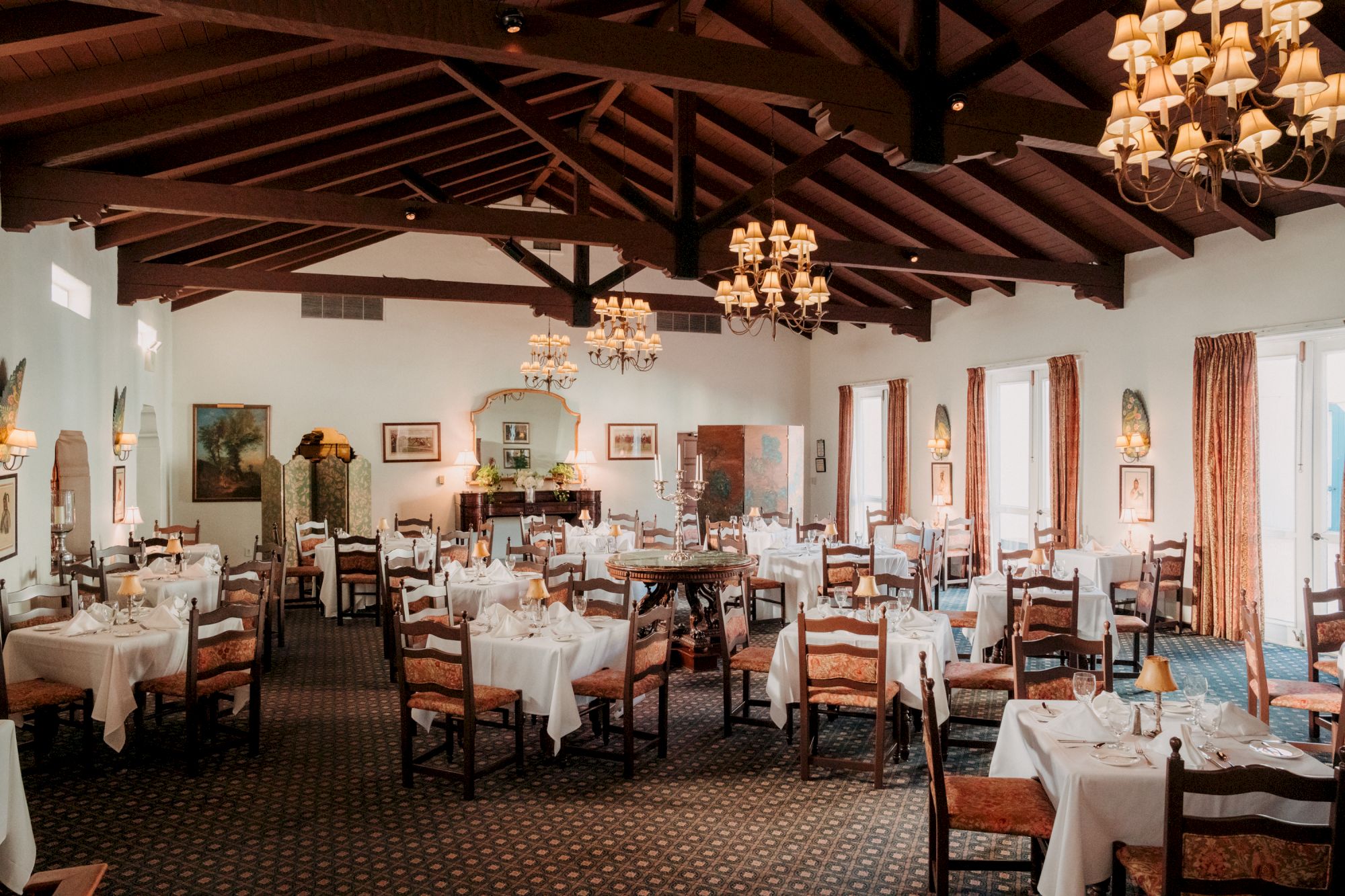 A banquet hall with wooden beams, chandeliers, and round tables set with white tablecloths and chairs, ready for dining, elegant and spacious.