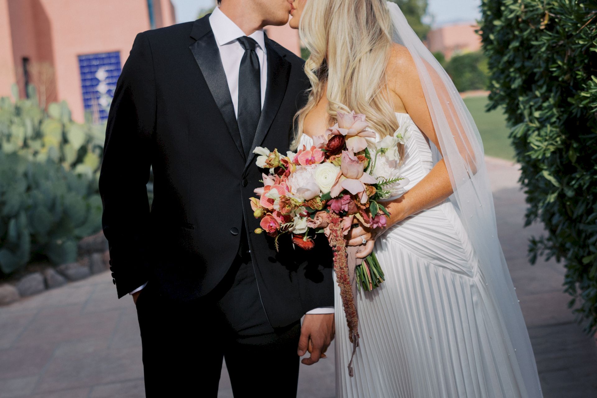 A groom in a suit and a bride in a white dress kiss outdoors, holding a colorful flower bouquet.