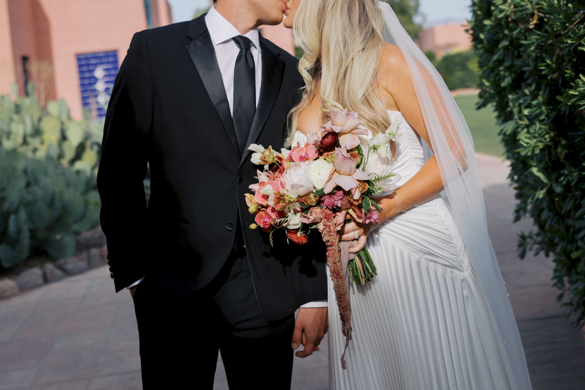 A groom in a suit and a bride in a white dress kiss outdoors, holding a colorful flower bouquet.