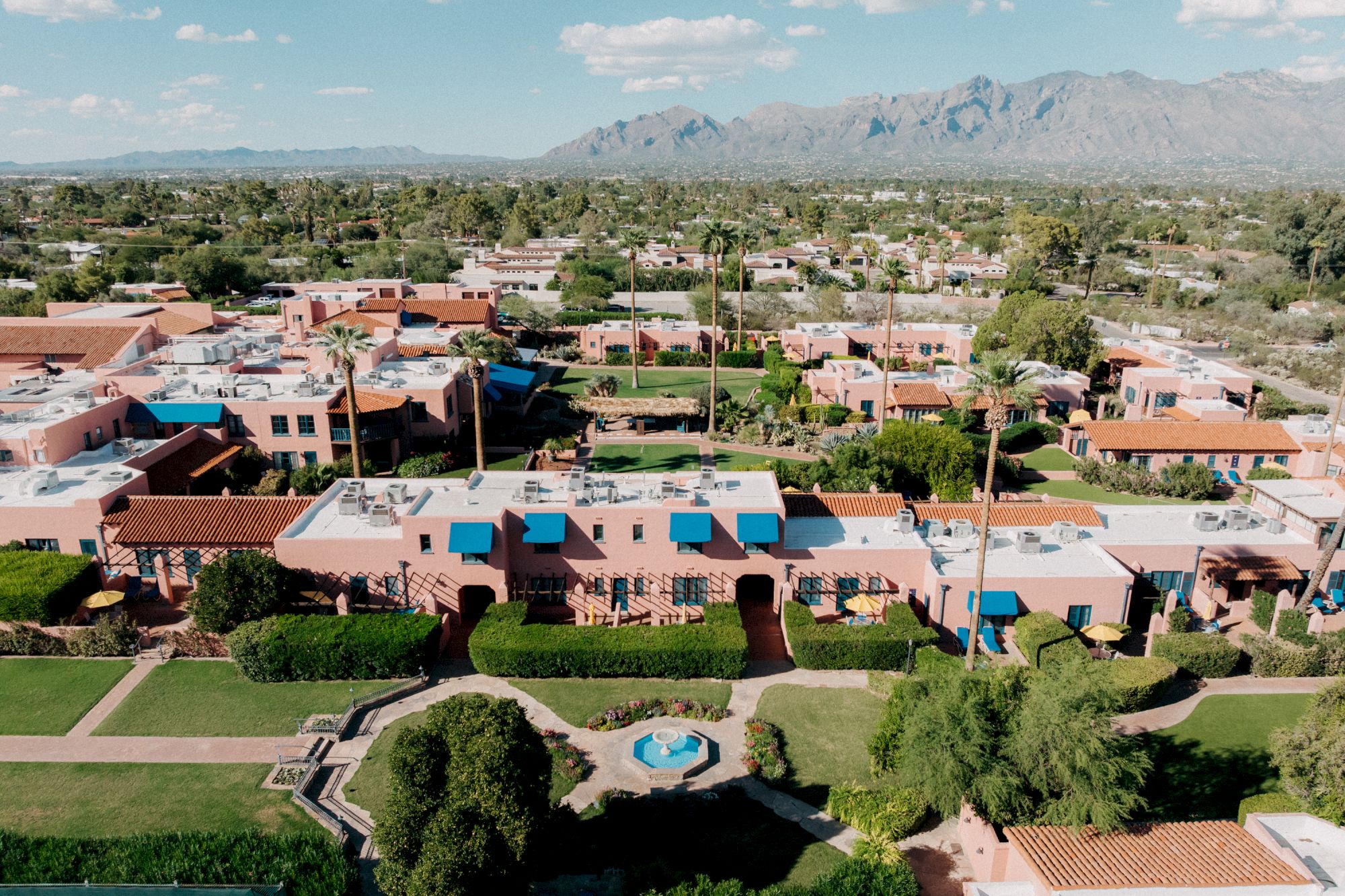 Aerial view of a suburban neighborhood with single-story houses, palm trees, and a central garden area; mountains in the distance, clear sky.