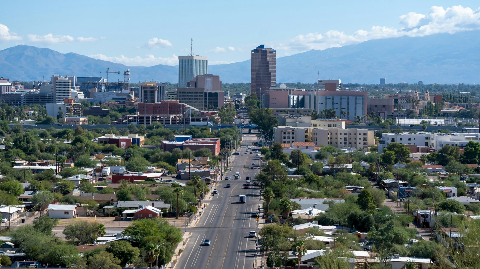 City skyline over a grid of low-rise homes and trees; a central road leads to taller buildings with mountains in the distance.