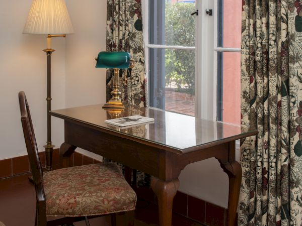 A classic wooden desk with a green banker lamp, a chair, a floor lamp, floral curtains, and a window letting light into a cozy study corner.