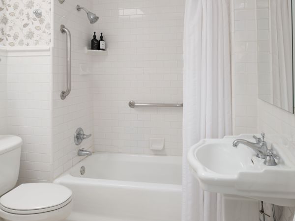 A clean white bathroom with a bathtub, shower curtain, grab bars, a small sink, and a toilet, all in a bright, compact layout.