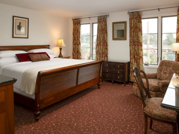 A cozy hotel room with a large wooden bed, lamps, patterned carpet, armchairs, a desk, and windows with floral curtains. Both bed and furniture look classic and comfortable.
