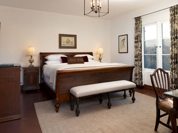 A traditional hotel room with a wooden king bed, matching nightstands, a bench at the foot, two lamps, and a window with floral curtains.