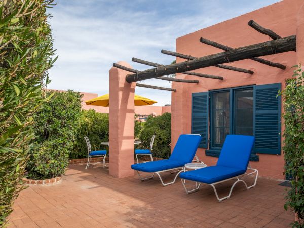 A sunny patio with pink walls, blue lounge chairs, wooden pergola beams, blue shutters, and potted greenery along a tiled terrace.