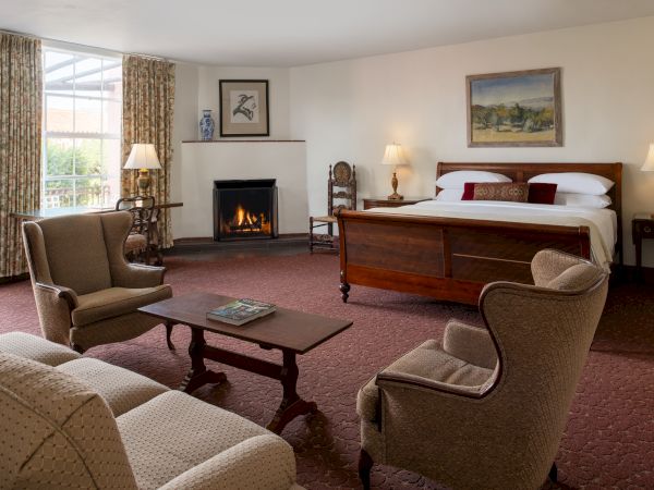 A cozy hotel suite with a fireplace, a large wooden bed, two armchairs, a coffee table, and soft lighting by lamps; curtains frame a sunlit window.