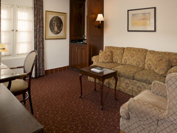 A cozy hotel living room with a patterned sofa, matching armchairs, a wooden coffee table, desk, lamp, and framed art on the walls.